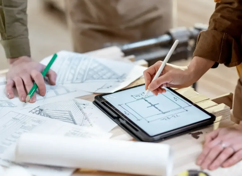 Hands on table covered with technical drawings with someone drawing on an ipad